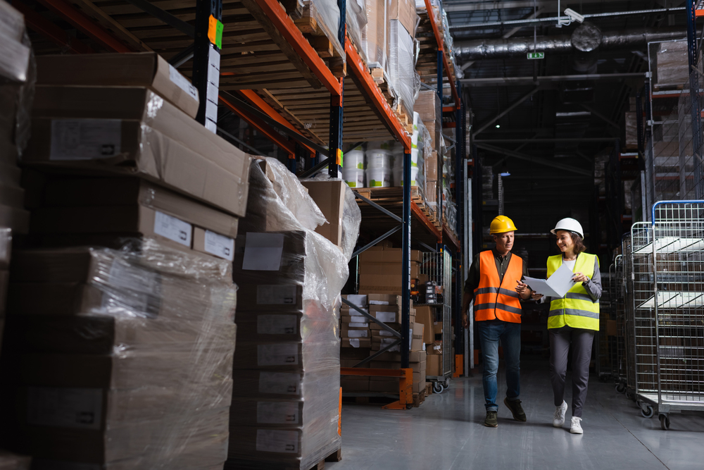 Two warehouse workers wearing high-visibility vests and helmets review fleet data while walking through an aisle lined with stacked pallets and inventory.